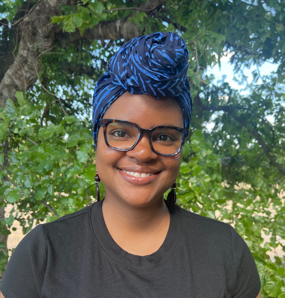 Photo of lab director standing in front of a tree wearing a black shirt, black earrings, square black and blue glasses, and a blue head wrap with a knot at the front.