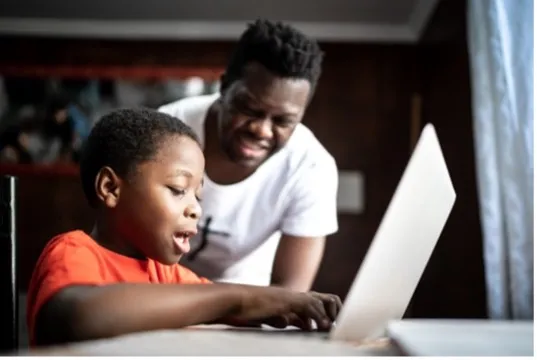 A young boy wearing an orange shirt sitting at a desk with a laptop with a man in a white shirt helping him